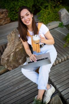 cute young woman drinking coffee sits on a bench with a laptop on the street.