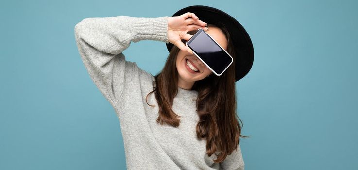 Pretty young smiling woman wearing black hat and grey sweater holding phone looking at camera isolated on background.Mock up, cutout, empty space