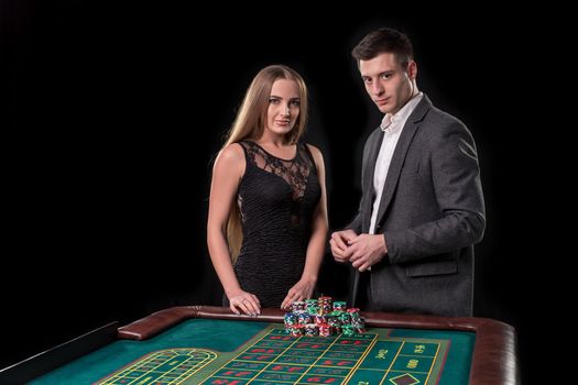 Elegant couple at the casino betting on the roulette, on a black background. A man in a suit with a beautiful young woman in a black dress