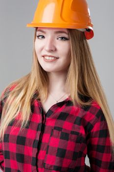 Portrait of a young attractive woman with blond hair in orange helmet and in a plaid shirt on a neutral gray background.