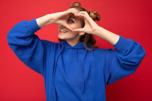 Photo of young positive happy smiling beautiful woman with sincere emotions wearing stylish clothes isolated over background with copy space and showing heart gesture with hands.