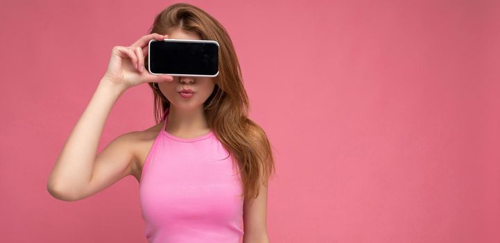 Photo of beautiful smiling young woman good looking wearing casual stylish outfit standing isolated on background with copy space holding smartphone showing phone in hand with empty screen display for mockup.