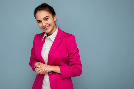 Portrait of young attractive beautiful positive smiling brunette woman with sincere emotions wearing casual white shirt and trendy pink jacket isolated over blue background with free space.