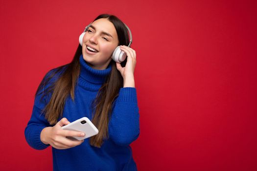 Photo of beautiful happy smiling young brunette woman wearing blue sweater isolated over red background wall using smartphone wearing white bluetooth headphones listening to cool music and enjoying. copy space