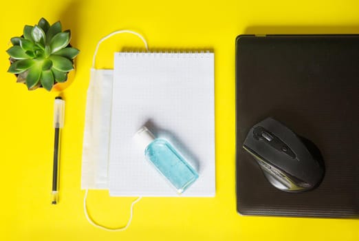 Top view of a desktop with notebooks, pens, and a laptop. Work from home using an online device, protection from coronavirus during the quarantine period. Protective masks and antiseptic on a yellow background.