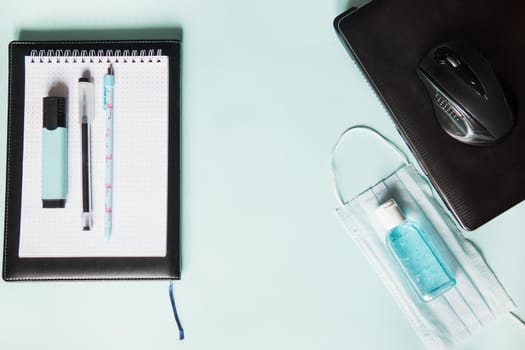 Top view of a desktop with notebooks, pens, and a laptop. Work from home using an online device, protection from coronavirus during the quarantine period. Protective masks and antiseptic on a mint background.