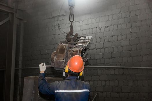 A working man in overalls, a protective helmet and a respirator controls heavy grinding equipment in the workshop of an industrial plant.