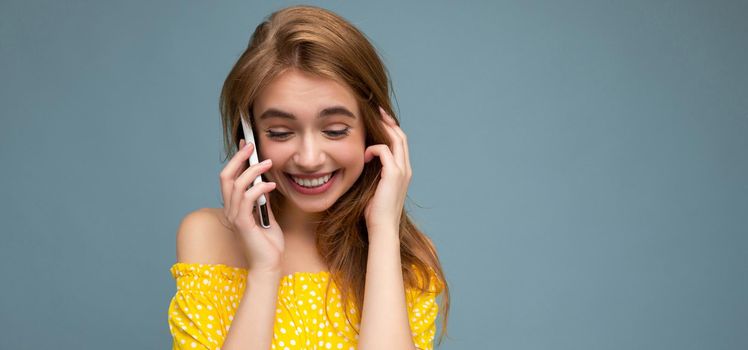 Attractive positive smiling young blonde woman wearing stylish yellow summer dress standing isolated over blue background holding and talking on mobile phone looking to down.