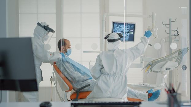 Professional dentistry team preparing for patient consultation using oral care equipment and modern technology. Stomatologists with ppe suits examining man during coronavirus epidemic