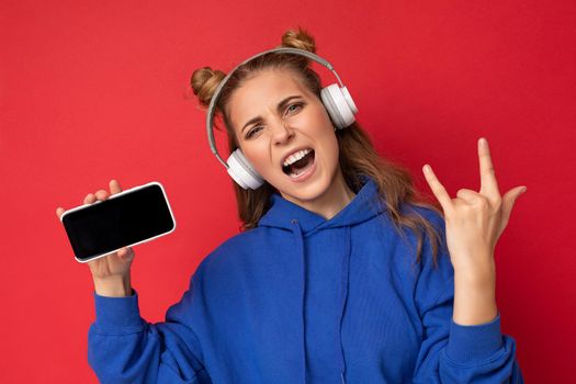 Emotional positive pretty young woman wearing stylish blue hoodie isolated over red background wearing white bluetooth wireless headphones and listening to music and showing mobile phone with empty space for cutout and showing rock and roll gesture.