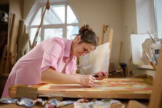 Cute woman paints on canvas in an art workshop. Artist creating picture. Art school or studio. Work with paints, brushes and easel. Hobby and leisure concept. Woman painter at workspace.
