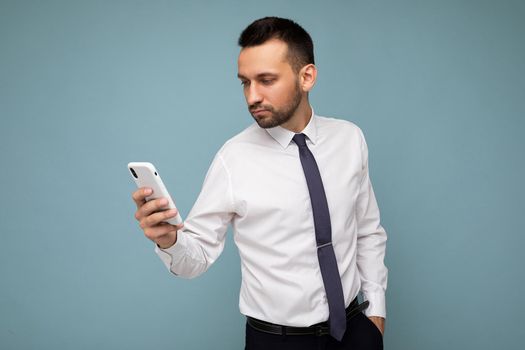 Photo shot of handsome concentrated good looking young businessman wearing casual outfit poising isolated on background with empty space holding in hand and using mobile phone messaging sms looking at smartphone display screen.