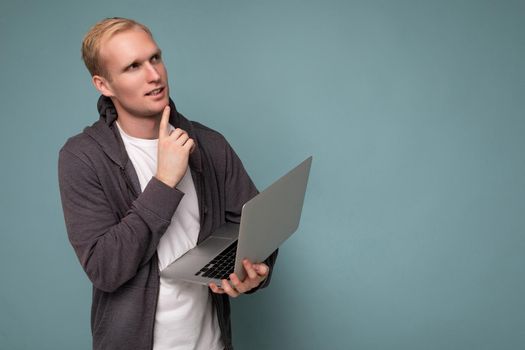 Side profile photo shot of handsome thoughtful blonde man holding computer laptop posing finger near chin wearing white t-shirt and grey sweater looking to the side isolated over blue background. Copy space