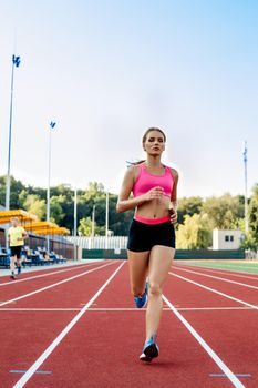 Sporty fitness woman jogging on red running track in stadium. Training summer outdoors on running track line with green trees on background. Sport, healthy lifestyle concept. Front view