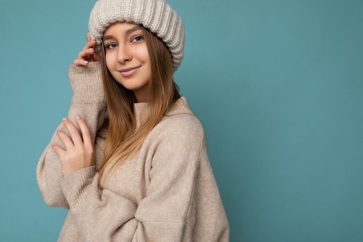 Portrait shot of attractive positive sexy cute adult dark blonde woman standing isolated on blue background wall wearing beige warm sweater and winter beige hat looking at camera. Copy space