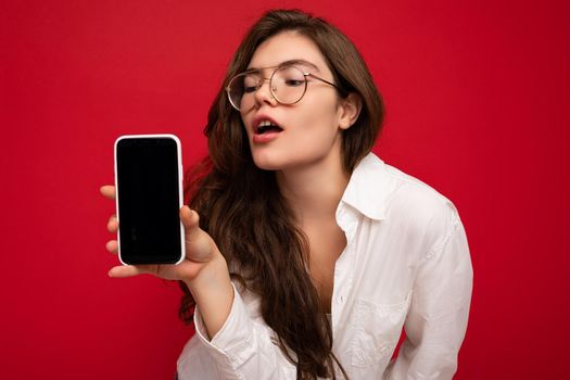photo of sexy beautiful young brunette woman wearing white shirt and optical glasses isolated over red background holding in hand and showing mobile phone with empty screen for cutout looking at gadjet screen.