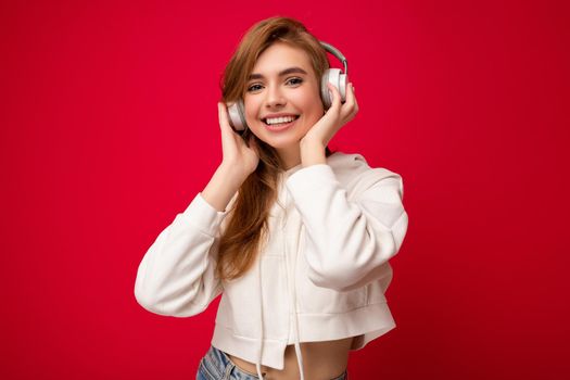 Photo of beautiful happy smiling young blonde woman wearing white hoodie isolated over colourful background wall wearing white wireless bluetooth earphones listening to cool music and enjoying looking at camera.