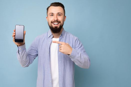 Photo of handsome smiling adult male person good looking wearing casual outfit standing isolated on background with copy space holding smartphone showing phone in hand with empty screen display for mockup pointing at gadjet looking at camera.