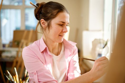 Cute woman paints on canvas in an art workshop. Artist creating picture. Art school or studio. Work with paints, brushes and easel. Hobby and leisure concept. Woman painter at workspace.