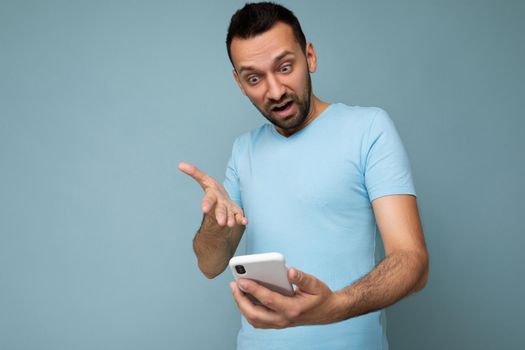 Photo of asking dissatisfied handsome young man with beard wearing everyday blue t-shirt isolated over blue background holding and using mobile phone communication online on the internet looking at gadjet display and having questions.