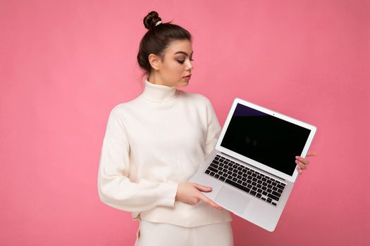 Photo of beautiful woman with gathered brunette hair wearing white sweater holding computer laptop and looking at the netbook isolated over pink wall background.
