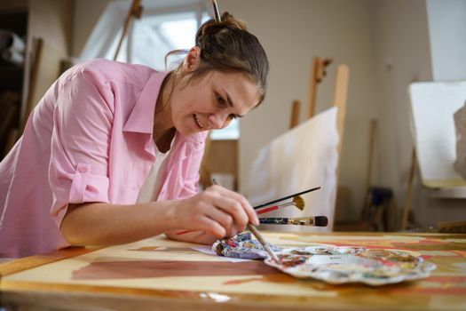 Cute woman paints on canvas in an art workshop. Artist creating picture. Art school or studio. Work with paints, brushes and easel. Hobby and leisure concept. Woman painter at workspace.