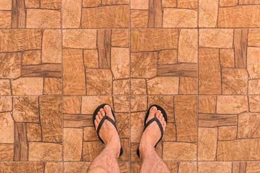 Legs of a man in black flip flops stand on a brown tiled floor with an abstract stone pattern background, top view.