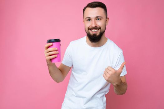 photo of handsome smiling young brunette unshaven male person with beard wearing white t-shirt isolated over pink backgroung wall holding paper coffee cup drinking and looking at camera and showing thumbs up gesture.