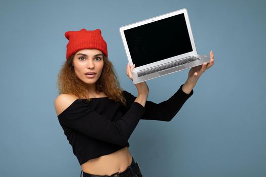 Beautiful amazed surprised shocked asking thoughtful young brunette curly woman wearing black crop top and red and orange do-rag isolated over light blue wall background holding computer laptop with empty screen monitor looking at camera. Mock up, copy space, empty space