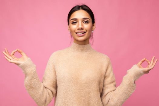 Portrait of young positive calm happy beautiful brunette woman with sincere emotions wearing casual beige jersey isolated over pink background with free space and keeping hands in mudra sign. Yoga concept.