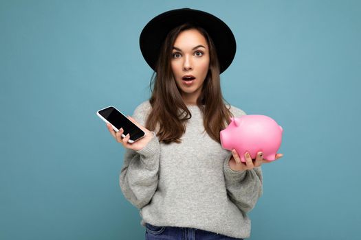 Portrait photo of shocked amazed young pretty nice woman with dark hair wearing stylish clothes and black hat isolated on blue background with copy space and holding pink pig money box and mobile phone.
