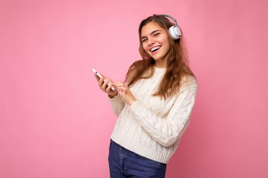 Photo shot of beautiful joyful smiling young female person wearing stylish casual outfit isolated over colorful background wall wearing white bluetooth wireless earphones and listening to music and using mobile phone looking at camera.