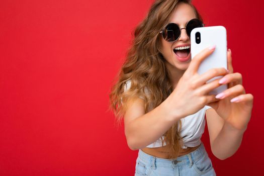 Closeup photo of amazing happy beautiful young blonde woman holding mobile phone taking selfie photo using smartphone camera wearing sunglasses everyday stylish outfit isolated over colorful wall background looking at device screen.