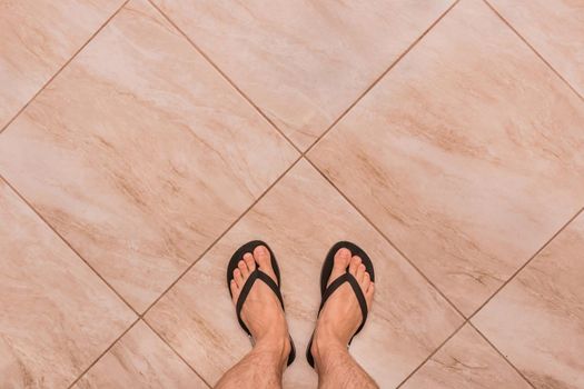 Legs of a man in black flip flops stand on a tiled floor background, top view.