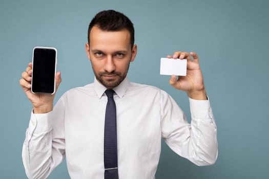 Closeup photo of handsome good looking brunet man with beard wearing casual white shirt and tie isolated on blue background with empty space holding in hand and showing mobile phone with empty screen for mockup and credit card looking at camera.