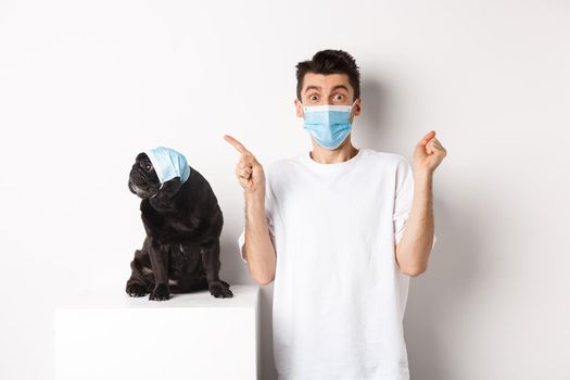 Covid-19, animals and quarantine concept. Young man and black dog wearing medical masks, pug looking at upper left corner and owner showing promo offer, rejoicing.