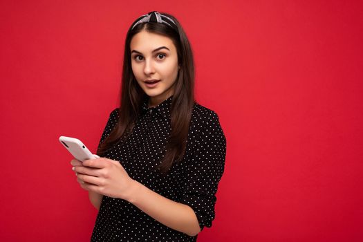 Photo of beautiful brunette girl standing isolated over red wall wearing casual stylish black clothes using mobile phone writing sms looking at camera. copy space