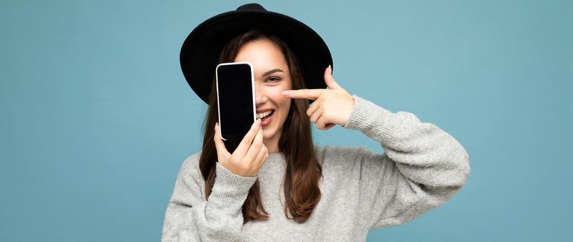 panoramic Photo of Beautiful positive woman wearing black hat and grey sweater holding mobilephone showing smartphone isolated on background looking at camera.Mock up, cutout, free space