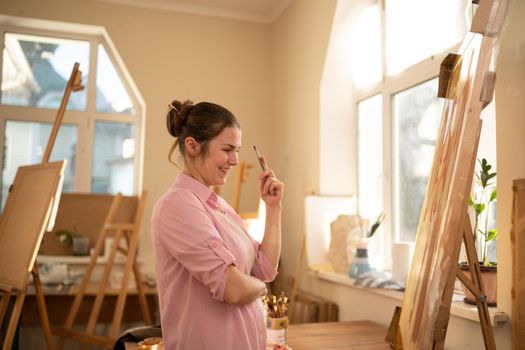 Cute woman paints on canvas in an art workshop. Artist creating picture. Art school or studio. Work with paints, brushes and easel. Hobby and leisure concept. Woman painter at workspace.