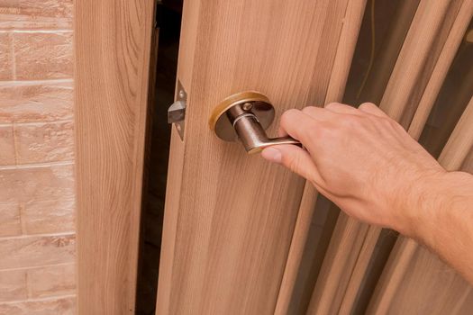 A man's hand holding an iron door handle opens or closes a wooden door indoors.