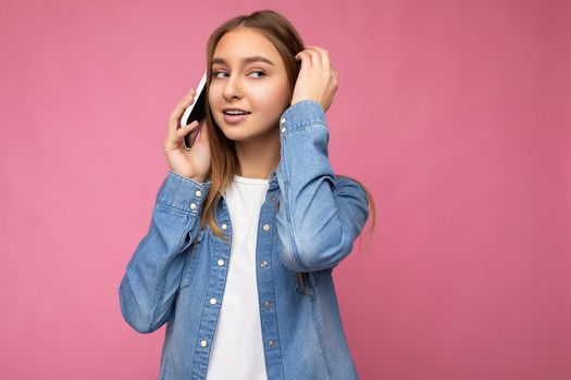 Photo of beautiful thoughtful young blonde woman wearing casual blue jean shirt isolated over pink background holding in hand and talking on mobile phone looking at camera and thinking.