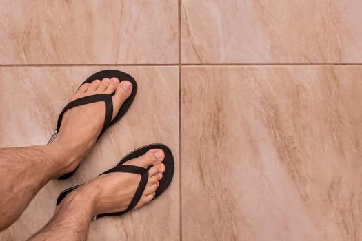 Legs of a man in black flip flops stand on a tiled floor texture background, top view.