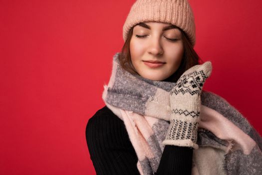 Photo of beautiful happy cute young brunette woman isolated over red background wall wearing winter scarf mittens and warm hat enjoying with close eyes.