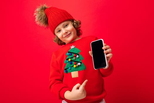 a young man with a smartphone in his hands against the background of a red wall, in a knitted hat and a sweater with a Christmas tree