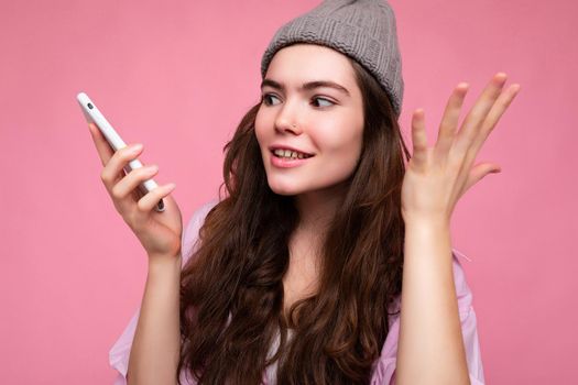 Closeup photo of Emotional attractive young brunette woman wearing stylish pink shirt and grey hat isolated over pink background holding in hand and using mobile phone communicating and recording voice message.