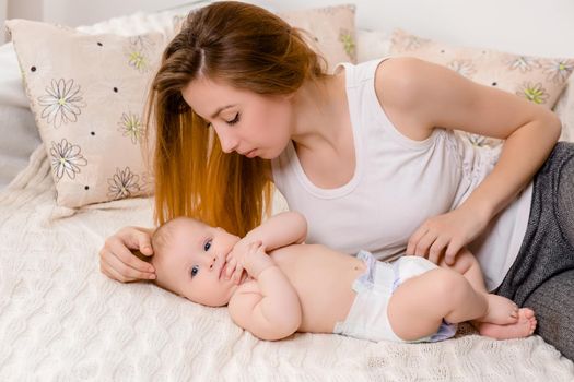 Mother and child on a white bed. Mom and baby girl in diaper playing in sunny bedroom. Parent and little kid relaxing at home. Family having fun together. Bedding and textile for infant nursery.