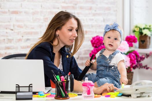 Working together is so fun Cheerful young beautiful businesswoman looking at telephone while sitting at her working place with her little daughter