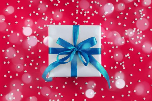 Gift box wrapped in white paper with a blue bow on festive crimson background with many snowflakes. Copyspace for your text. Flat lay style. Christmas, New Year or birthday celebration concept