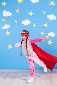 Six year blonde girl dressed like superhero having fun at home. Kid on the background of bright blue wall with white clouds and yellow stars. Girl power concept.