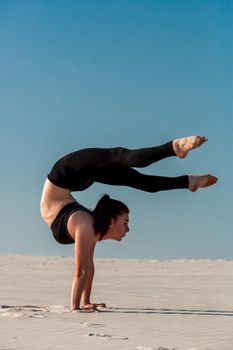 Young woman practicing inversion balancing yoga pose handstand on beach with white sand and bright blue sky